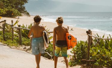 Two young surfers walk along the tropical beach path towards the ocean, carrying surfboards on a bright day.