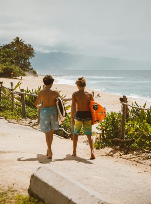 Two young surfers walk along the tropical beach path towards the ocean, carrying surfboards on a bright day.