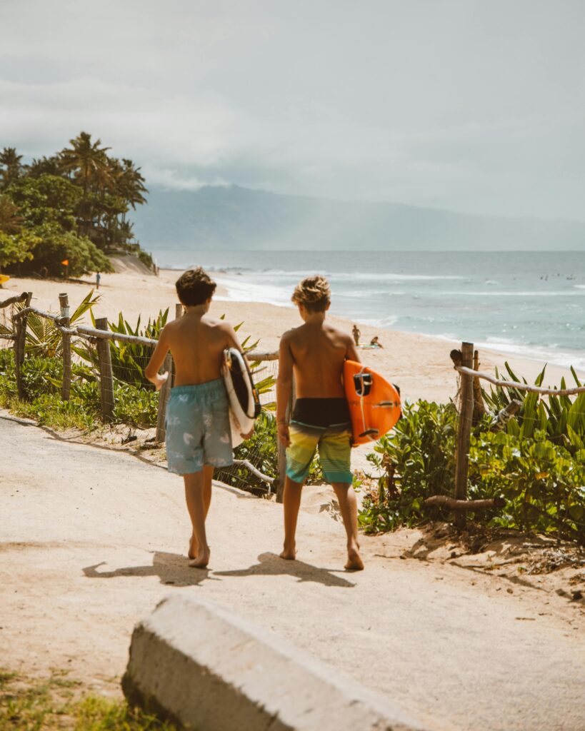 Two young surfers walk along the tropical beach path towards the ocean, carrying surfboards on a bright day.